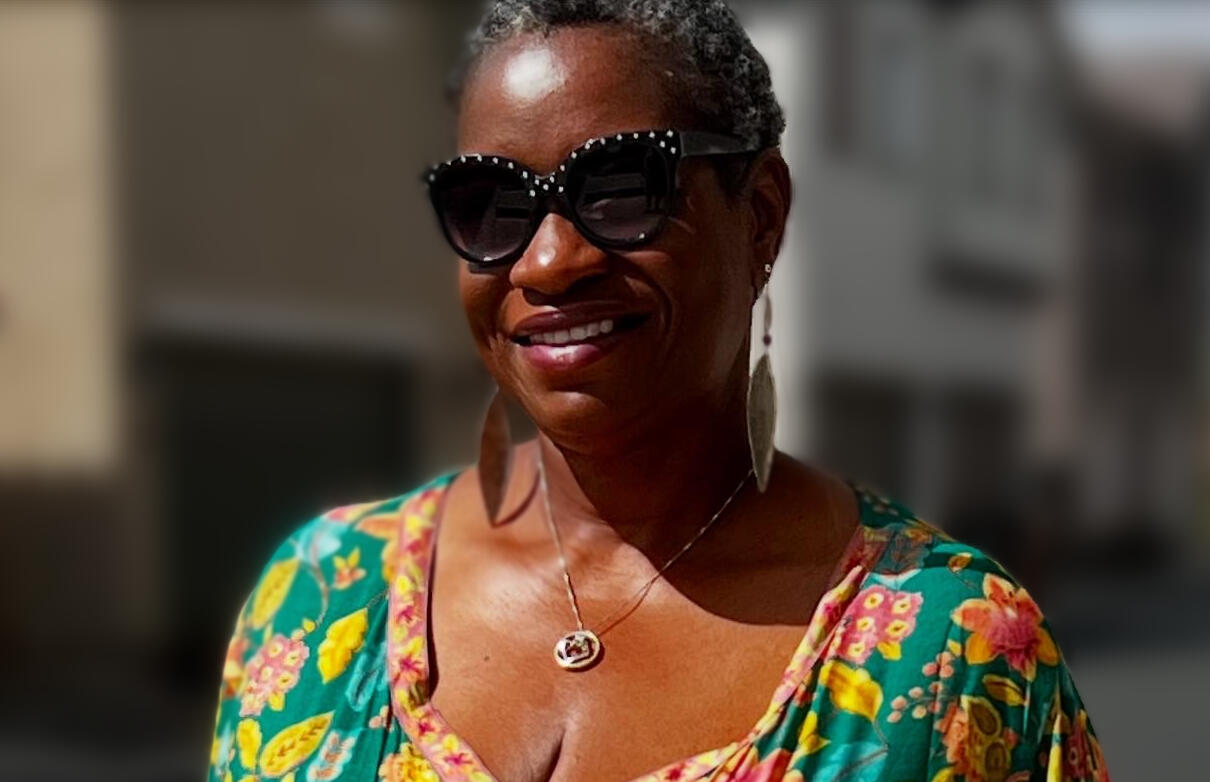 Kristina Daniele A smiling Black woman with short gray hair wearing large black sunglasses with decorative studs or rhinestones along the frames. She's dressed in a vibrant floral-patterned top or dress featuring bright colors including teal, yellow, pink, and green flower