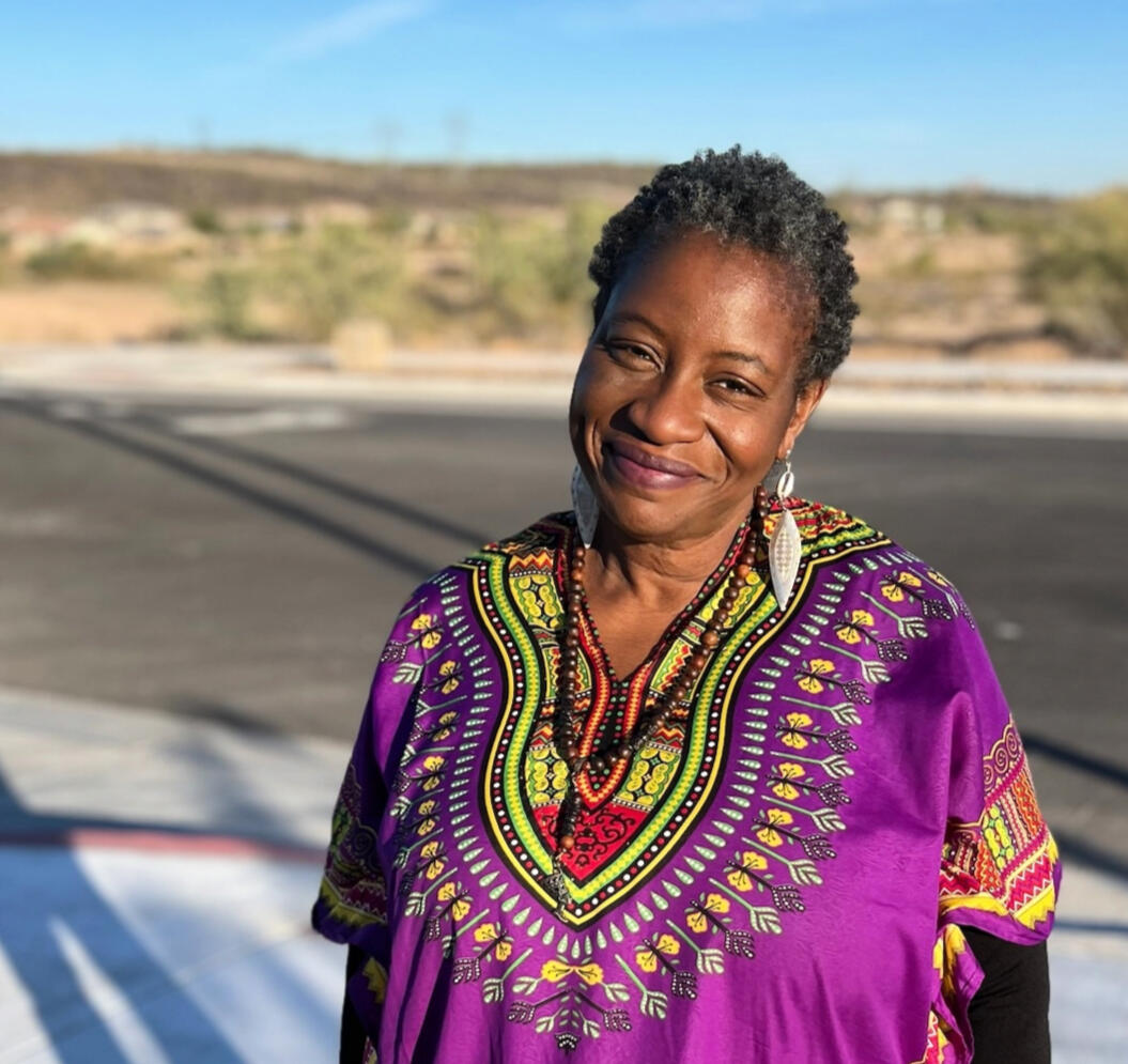 Kristina Daniele A smiling Black woman with short gray hair wearing large black sunglasses with decorative studs or rhinestones along the frames. She's dressed in a vibrant floral-patterned top or dress featuring bright colors including teal, yellow, pink, and green flower