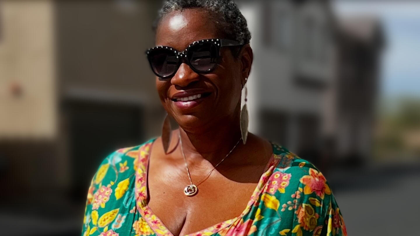 Kristina-Brooke Daniele A smiling Black woman with short gray hair wearing large black sunglasses with decorative studs or rhinestones along the frames. She's dressed in a vibrant floral-patterned top or dress featuring bright colors including teal, yellow, pink, and green flower