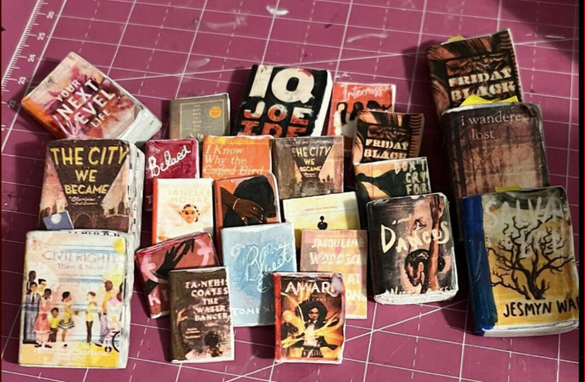 Kristina-Brooke Daniele A smiling Black woman with short gray hair wearing large black sunglasses with decorative studs or rhinestones along the frames. She's dressed in a vibrant floral-patterned top or dress featuring bright colors including teal, yellow, pink, and green flower